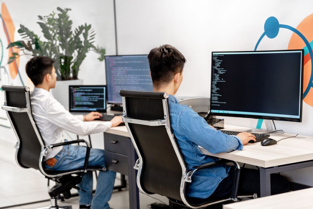 Services Two male developers at desks programming in a modern office workspace with large monitors.